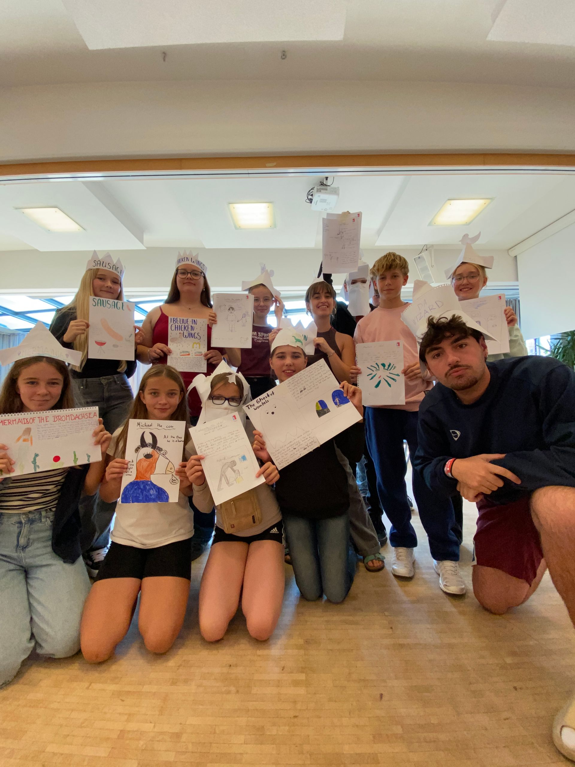 Eine Gruppe Kinder und ein Mann in einem Innenraum halten Kunstwerke in den Händen und tragen Papierkronen.