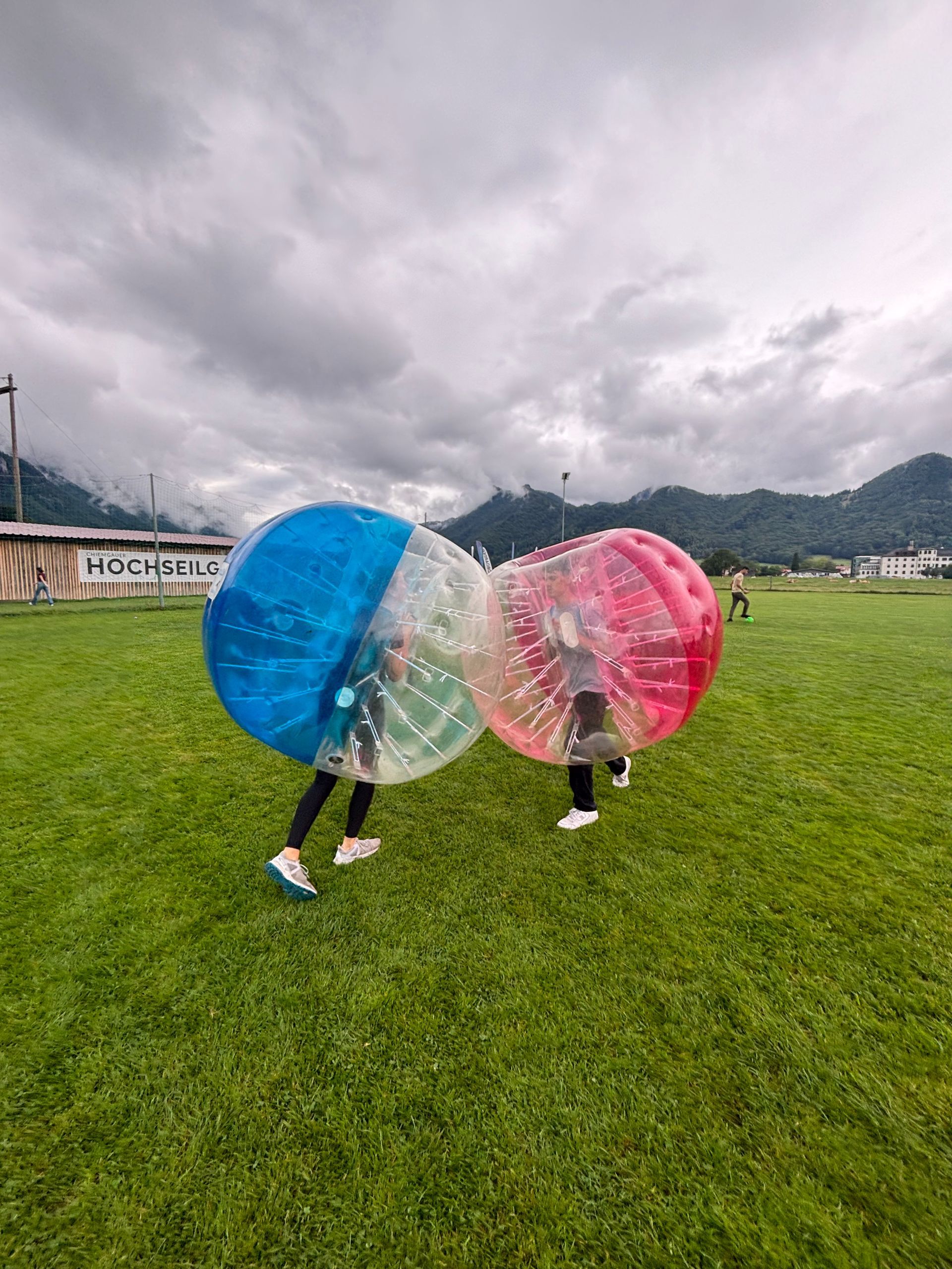 Zwei Personen in Schutzanzügen stoßen auf einer grünen Wiese zusammen. Im Hintergrund sind Berge und ein bewölkter Himmel zu sehen.