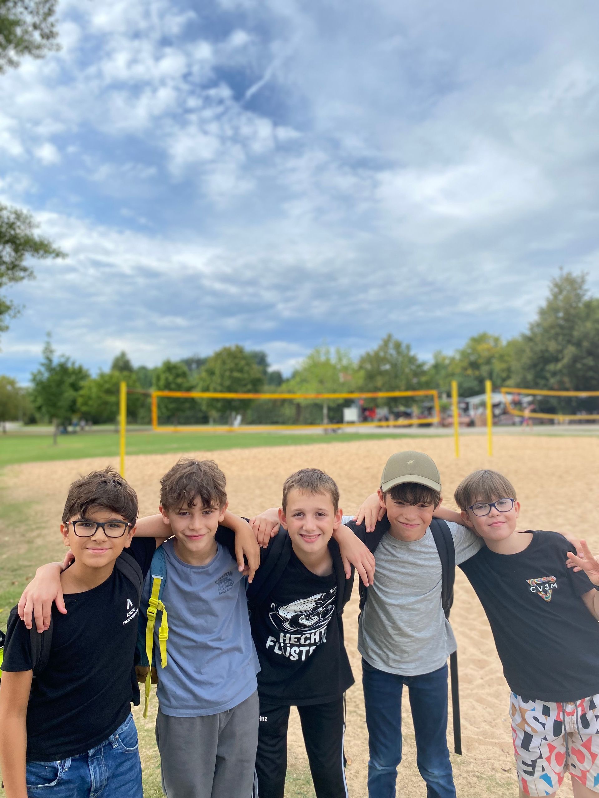 Fünf Jungen posieren mit umschlungenen Armen vor einem Volleyballnetz auf einem Sandplatz. Bewölkter Himmel.