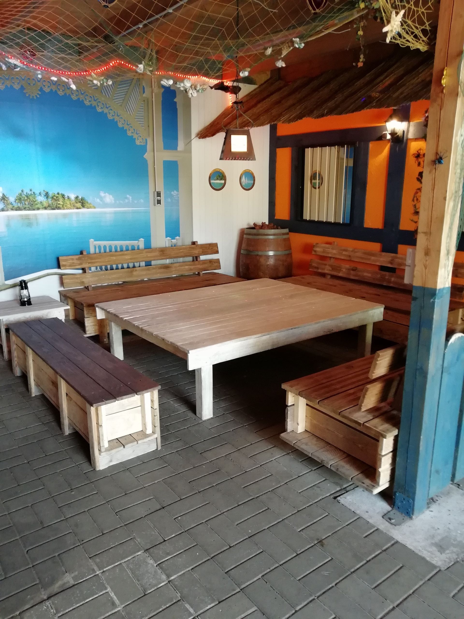 Sitzbereich im Freien mit Holztischen, Bänken und einem Wandgemälde mit Meeresmotiven.