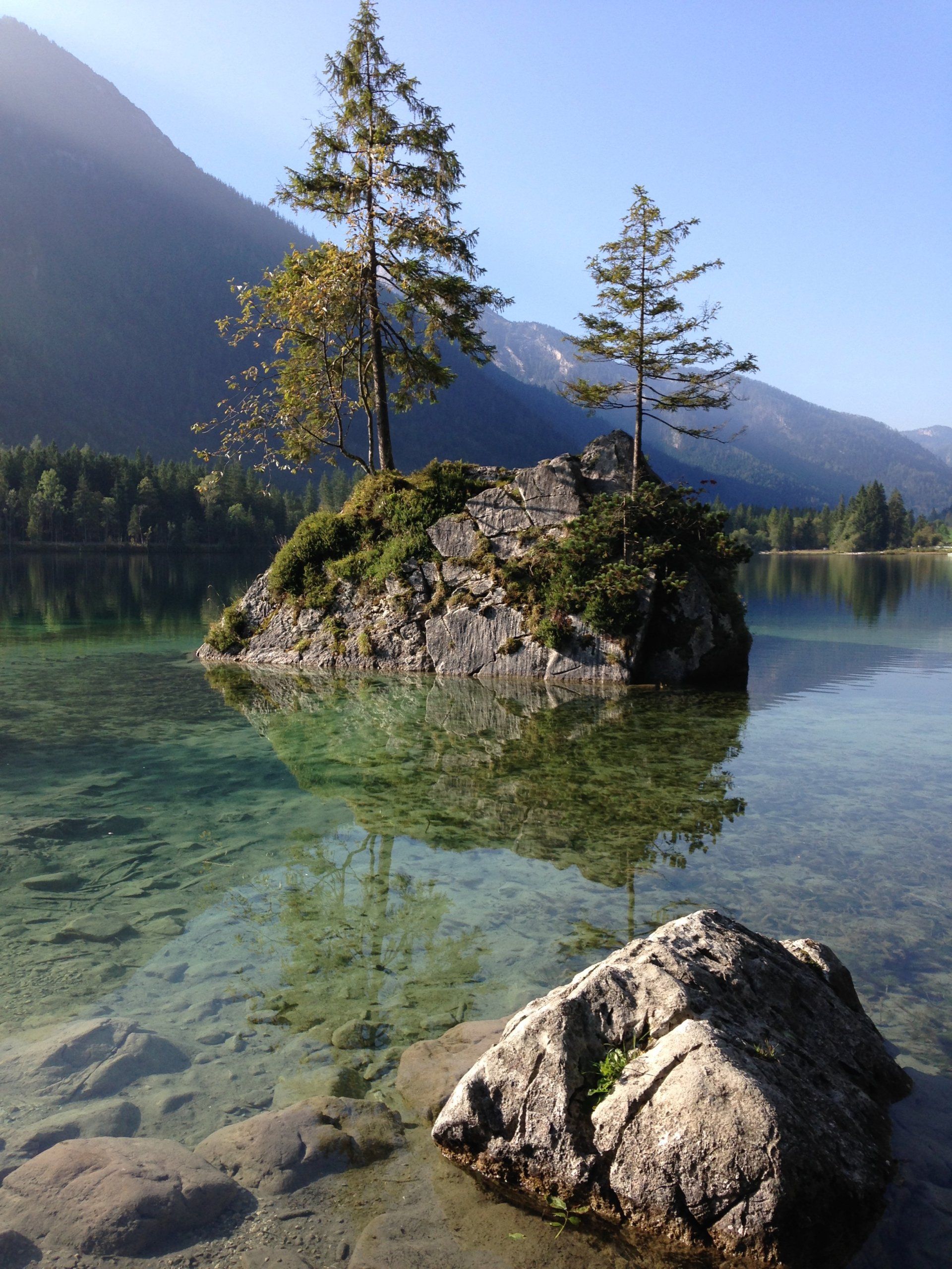 Insel mit Bäumen in einem klaren See, Berge im Hintergrund. Sonnenlicht spiegelt sich.
