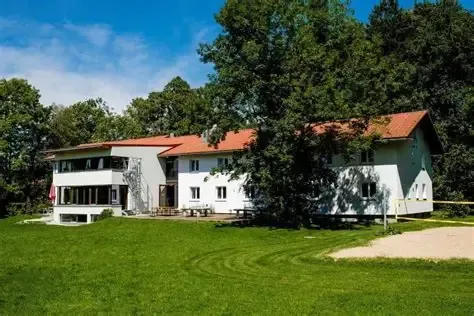 Weißes zweistöckiges Gebäude mit rotem Dach, auf einem grünen Rasen mit Bäumen im Hintergrund, unter blauem Himmel.