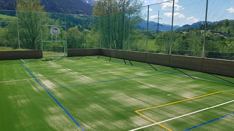 Grüner Sportplatz mit Basketballkorb, Fußballtor und umlaufendem Zaun vor einer Bergkulisse.