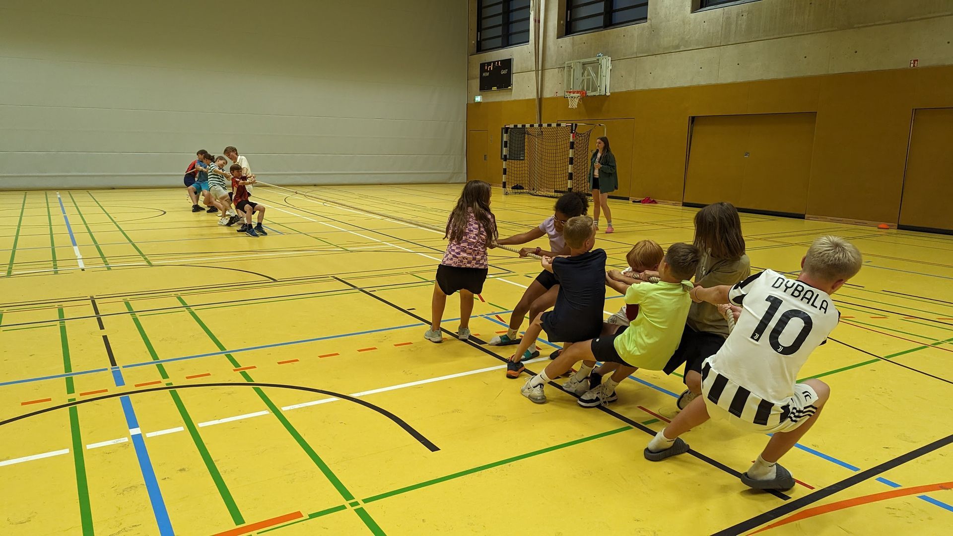 Kinder spielen Tauziehen in einer Turnhalle. Sie ziehen an einem Seil über einem gelben Boden.
