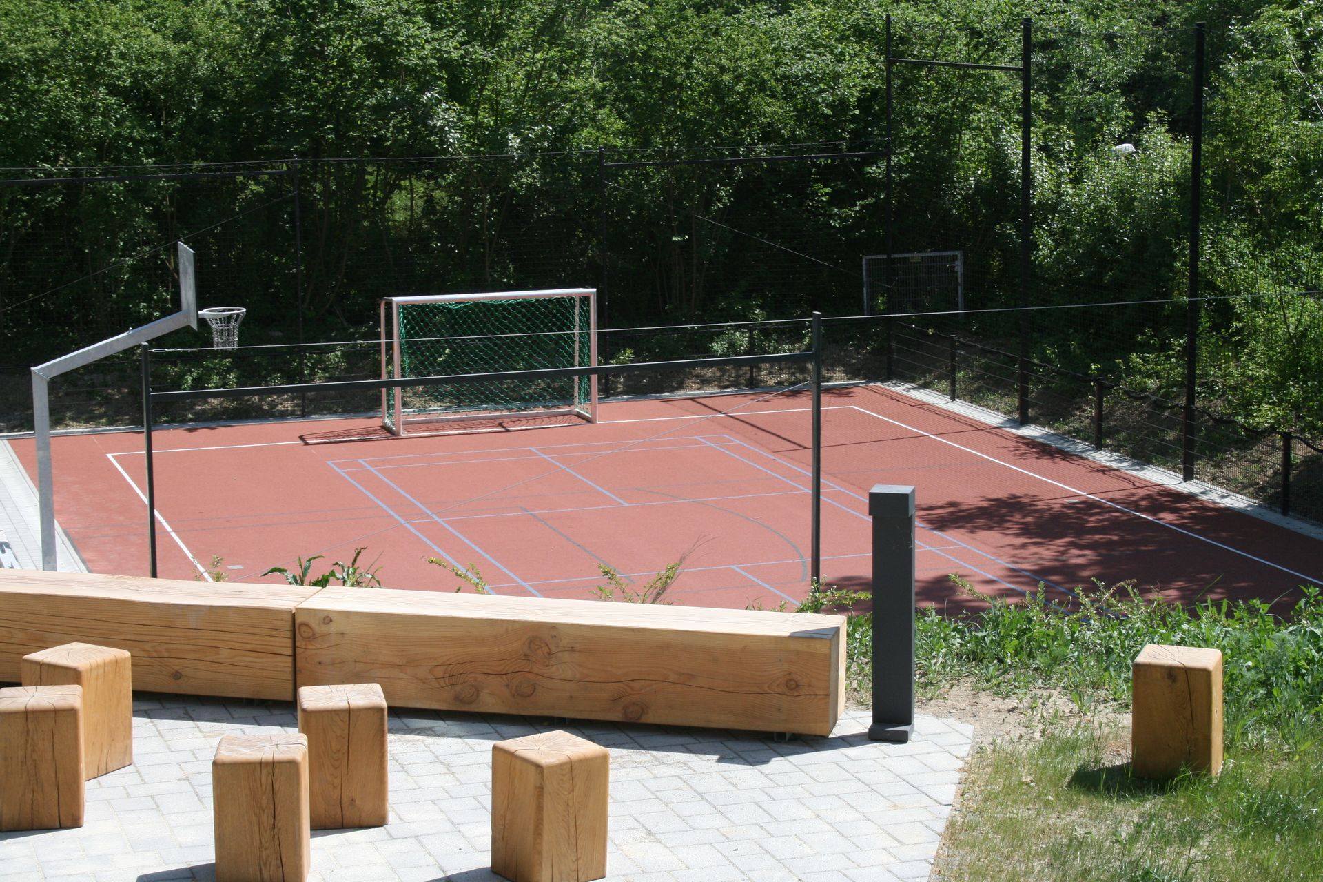Ein roter Sportplatz im Freien mit Basketballkörben und einem Fußballtor, umgeben von Bäumen und einer Holzbank.