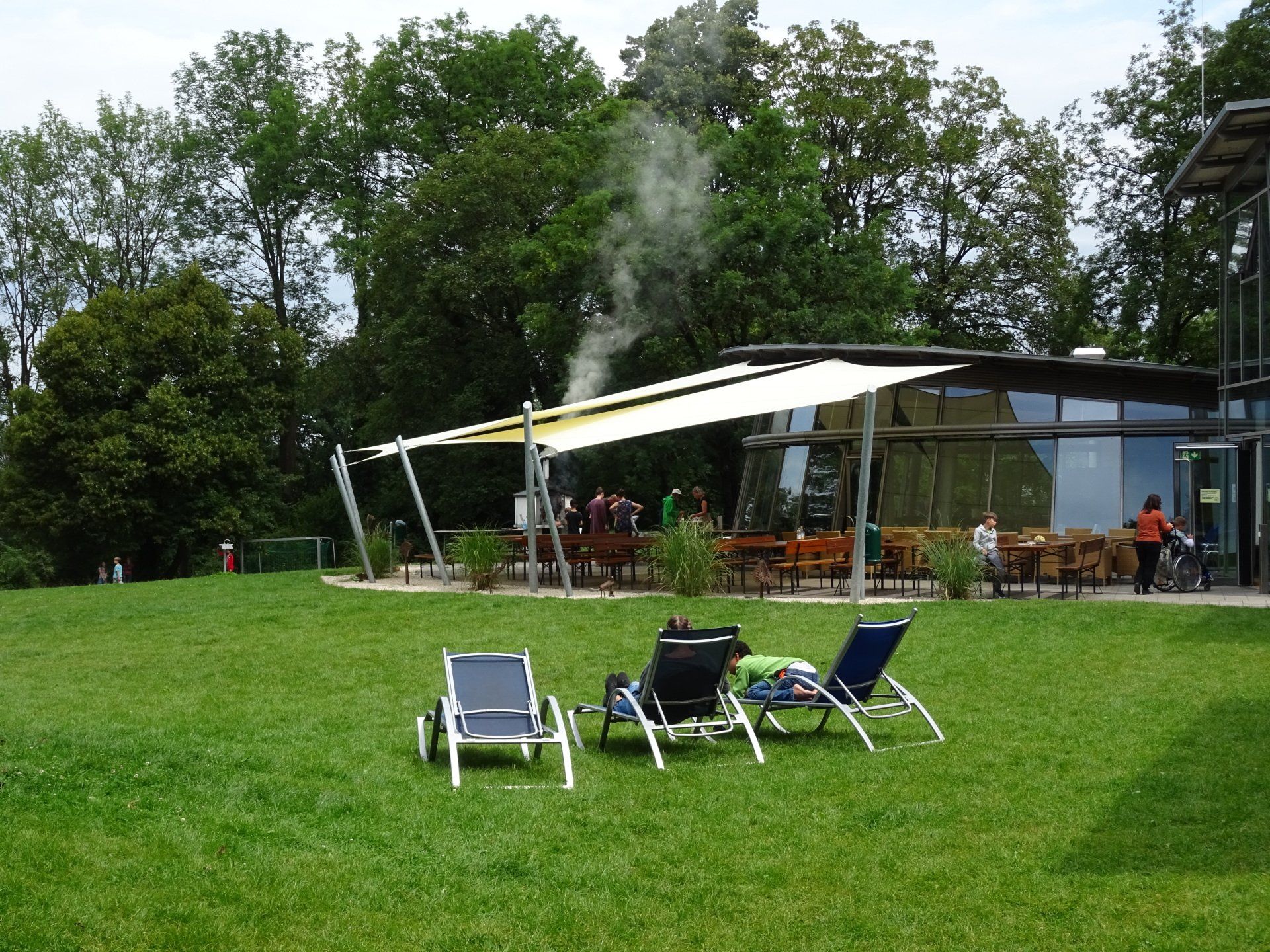 Liegestühle auf dem Rasen vor der Terrasse eines Restaurants, umgeben von Menschen, Bäumen und Rauch.