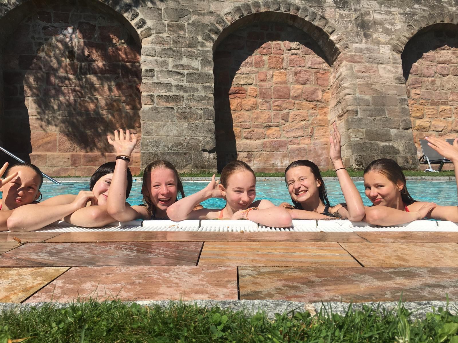 Sechs Personen in einem Schwimmbecken winken in der Nähe eines Steinbauwerks mit gewölbten Öffnungen.