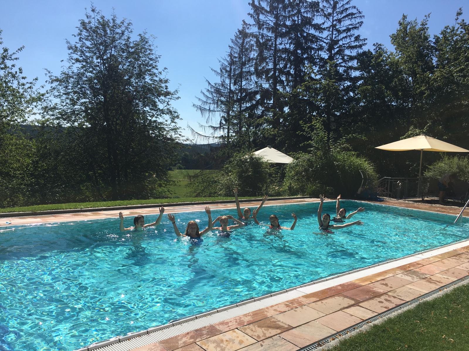 Menschen, die an einem sonnigen Tag in einem Schwimmbecken die Arme ausstrecken; im Hintergrund sind Bäume und Sonnenschirme zu sehen.