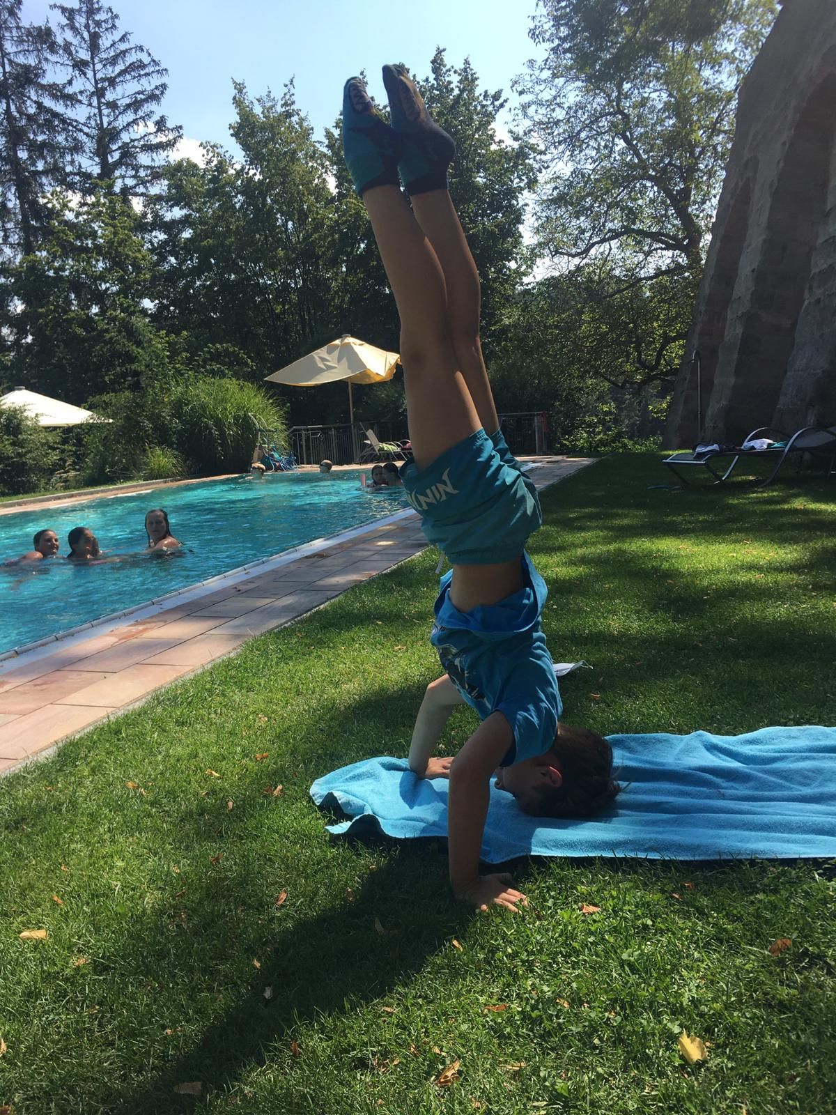 Eine Person macht einen Handstand auf einer Rasenfläche neben einem Pool.
