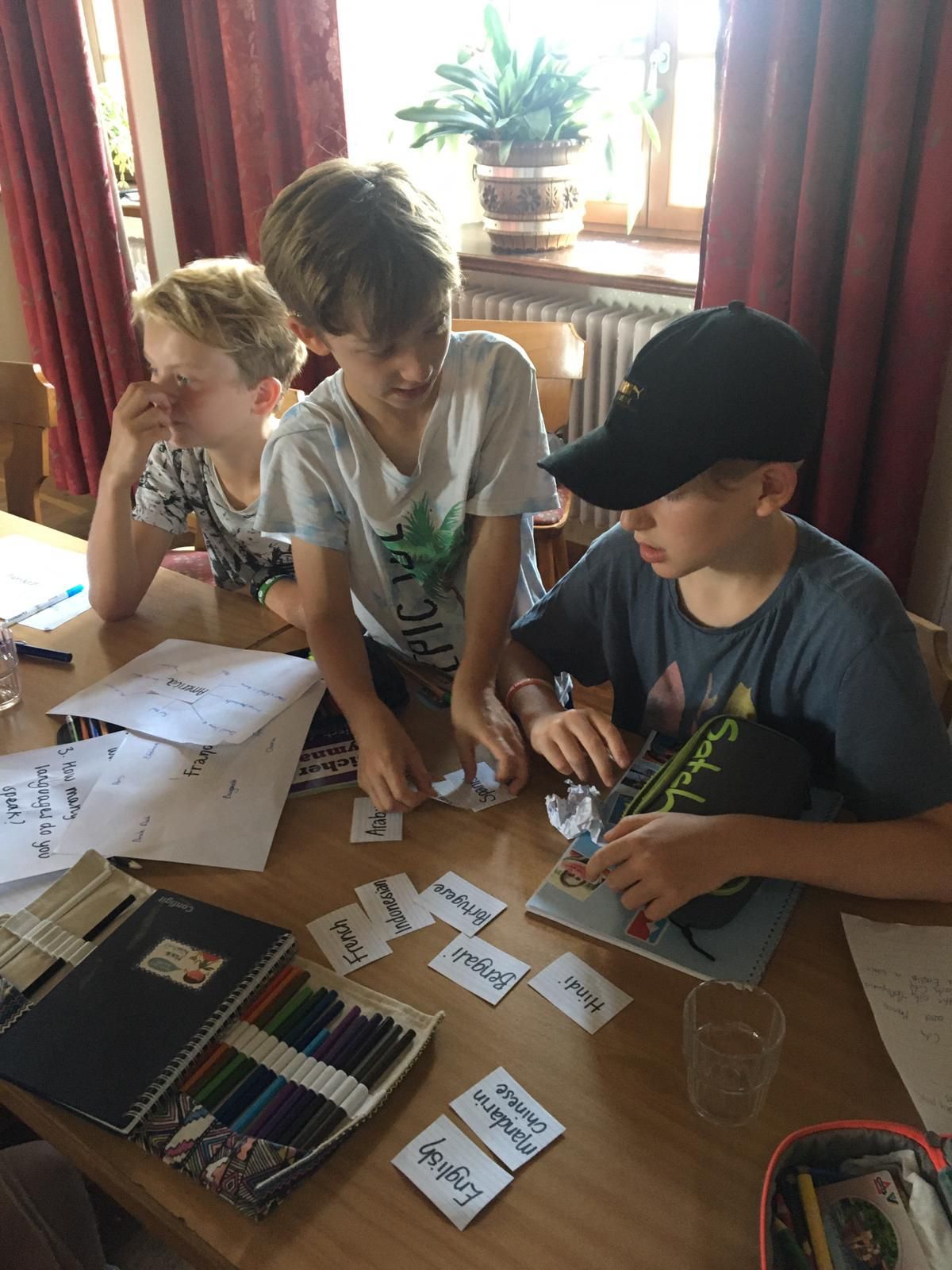 Drei Jungen sitzen an einem Tisch und studieren Aufsätze und Wortkarten. Im Hintergrund sind ein Fenster und Bücher zu sehen.