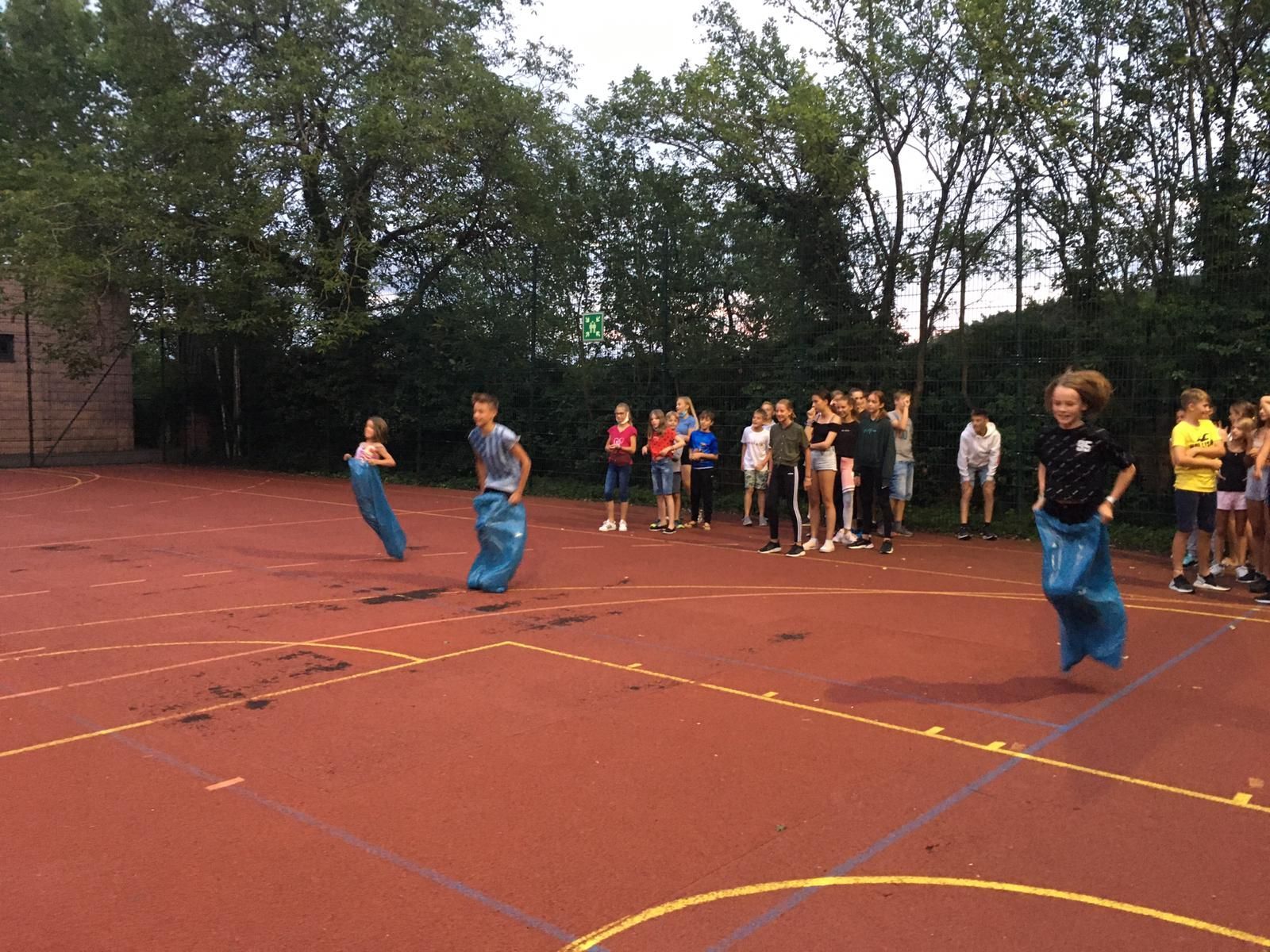 Kinder in Säcken nehmen an einem Sackhüpfen auf einem roten Sportplatz teil; Zuschauer beobachten die Szene.