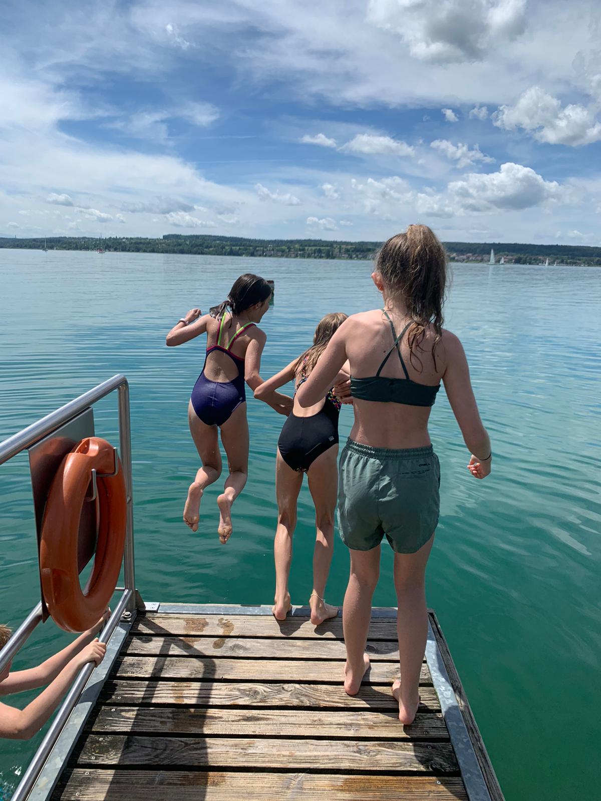 Drei Personen springen an einem sonnigen Tag von einem Steg in türkisfarbenes Wasser.