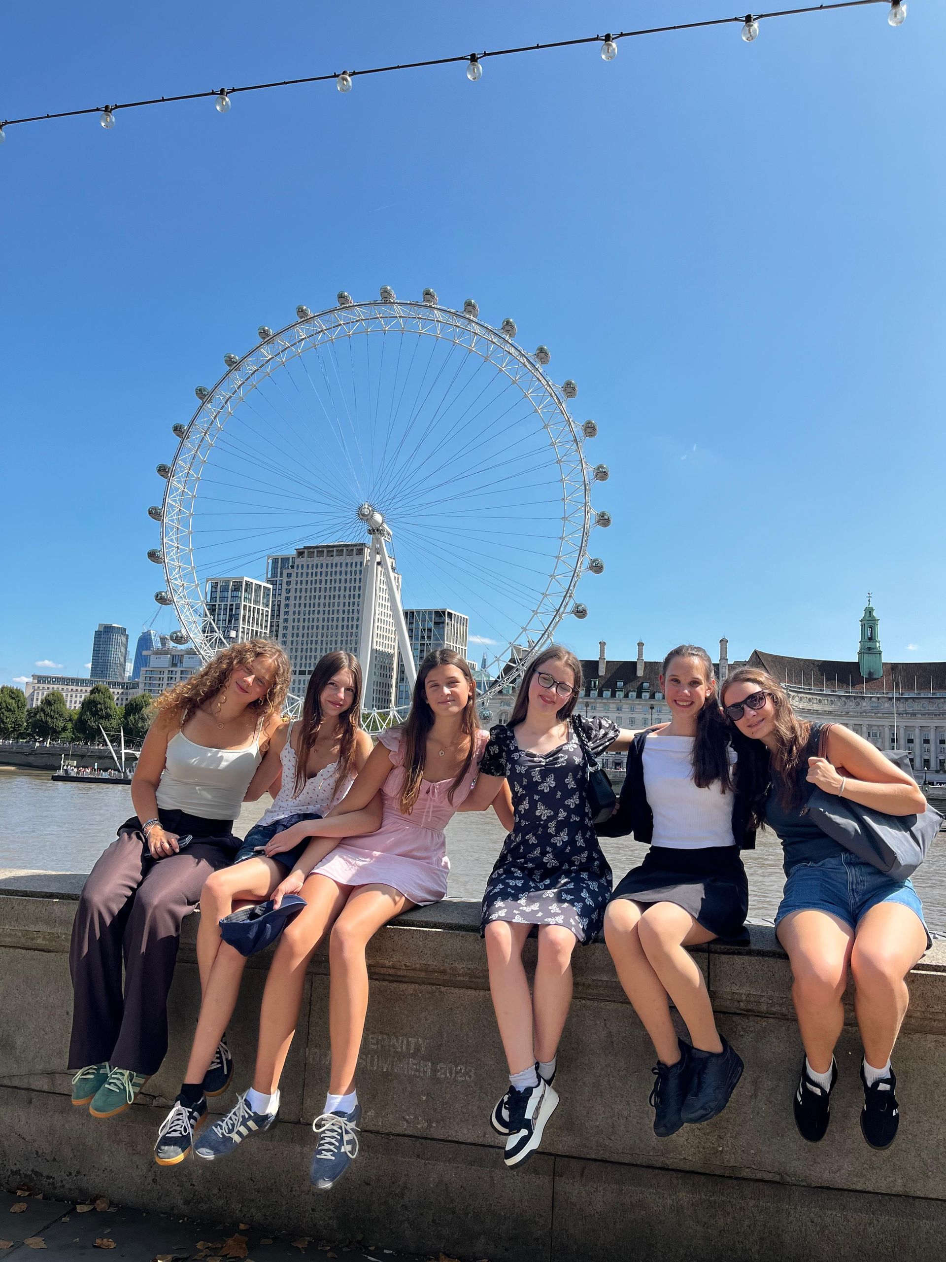 Sechs Personen sitzen an einem sonnigen Tag auf einer Mauer, im Hintergrund das London Eye.