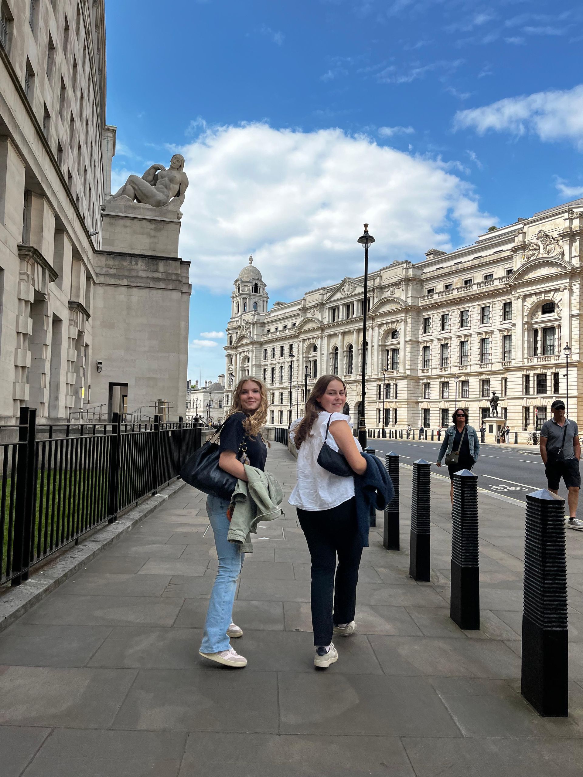 Zwei Personen gehen unter blauem Himmel auf einem Bürgersteig vor reich verzierten Gebäuden.
