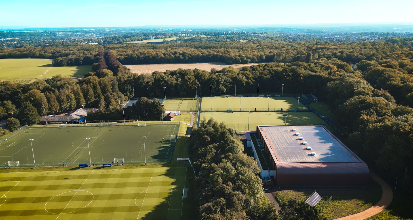 Luftaufnahme eines Sportkomplexes mit mehreren grünen Spielfeldern, Bäumen und einem großen braunen Gebäude.