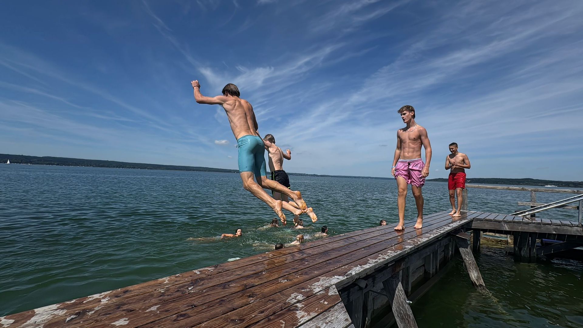 Menschen springen an einem sonnigen Tag von einem rostigen Pier ins Wasser. Blauer Himmel, Wasser und Badebekleidung.