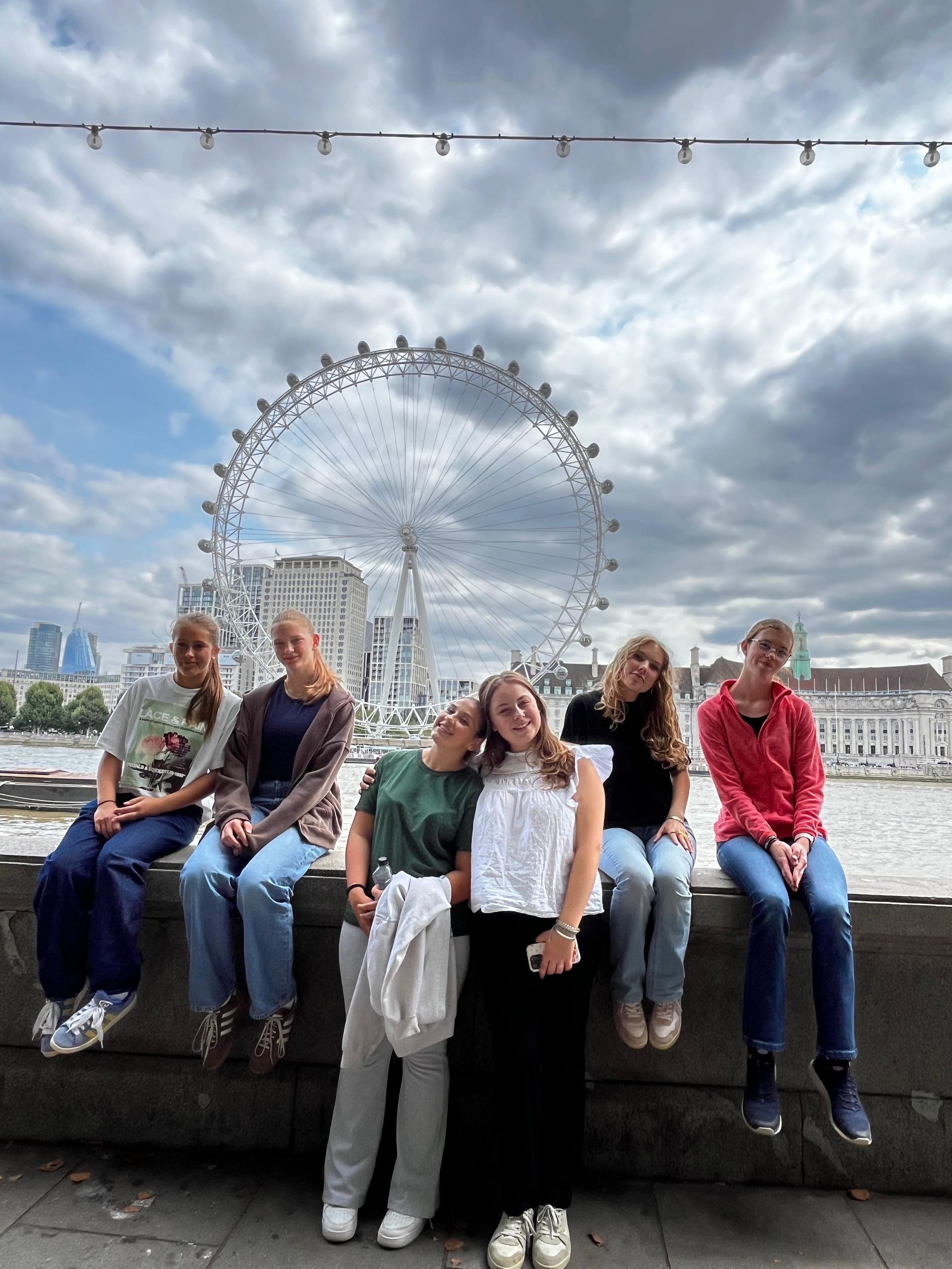 Sechs Personen sitzen auf einer Mauer, im Hintergrund das London Eye. Bewölkter Himmel, Fluss.