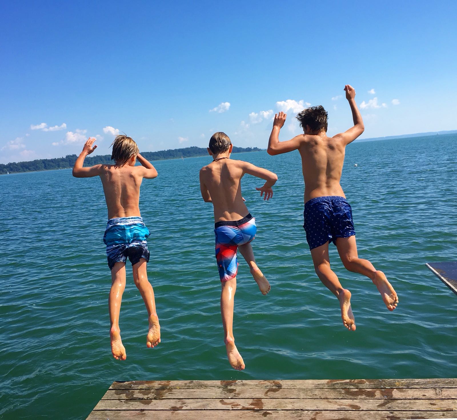 Drei Jungen springen an einem sonnigen Tag von einem Holzsteg ins Wasser.