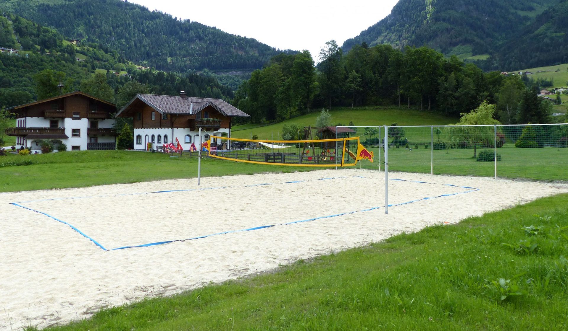 Beachvolleyballfeld mit Netz, umgeben von Gras und Bergen. Zwei Häuser im Hintergrund.