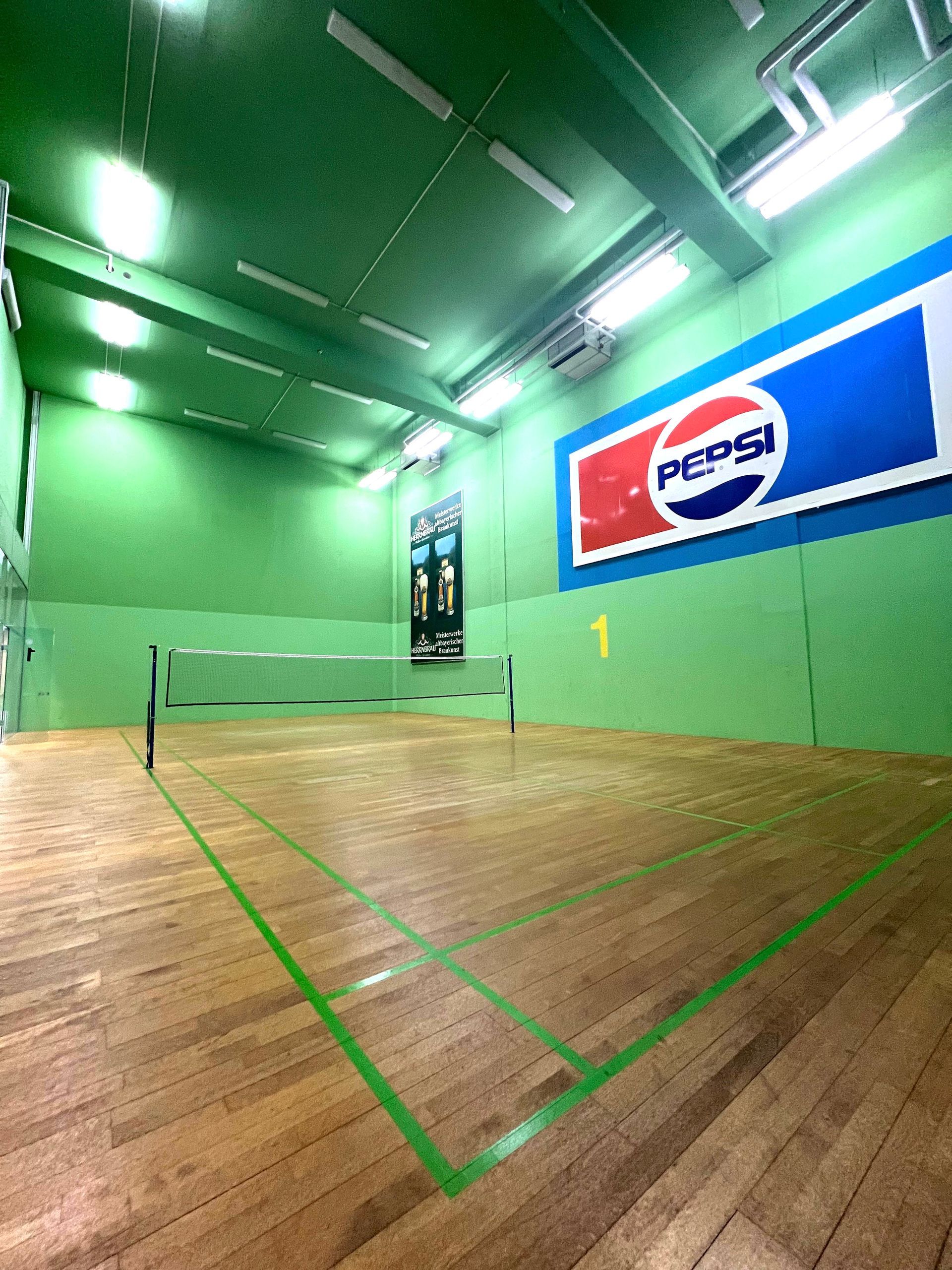 Badmintonfeld mit grünen Wänden, Holzboden, Netz und Pepsi-Logo.