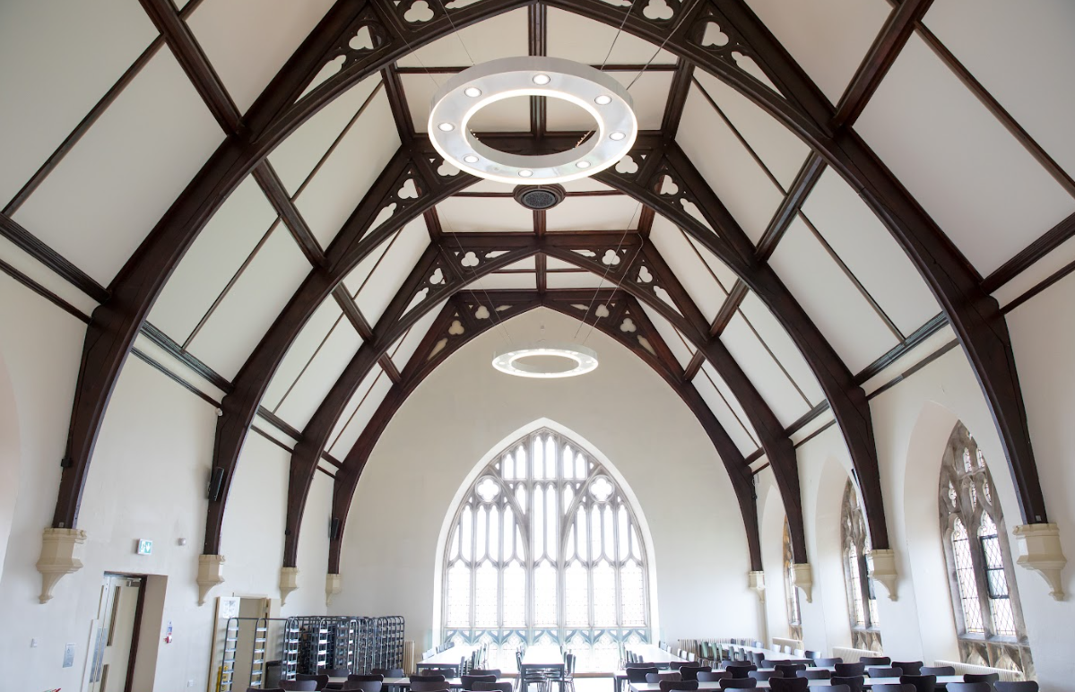 Innenansicht eines Gebäudes mit hoher, gewölbter Holzdecke, verzierten Fenstern und runden Leuchten.