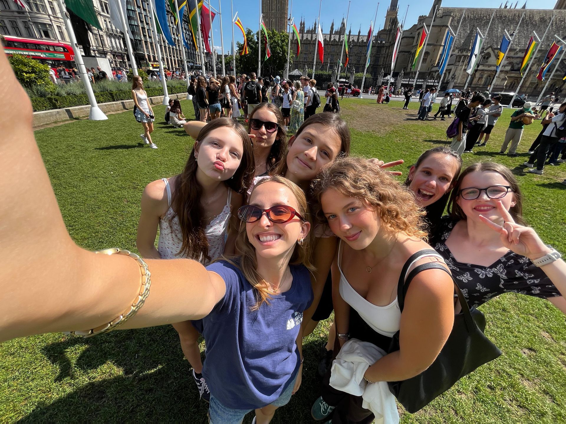 Eine Gruppe junger Leute macht im Freien ein Selfie; im Hintergrund sind Flaggen und ein Gebäude zu sehen.