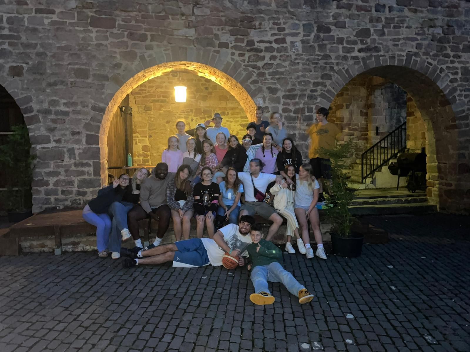 Eine Gruppe von Menschen posiert im Freien vor steinernen Torbögen. Einige sitzen, andere liegen auf dem Boden.