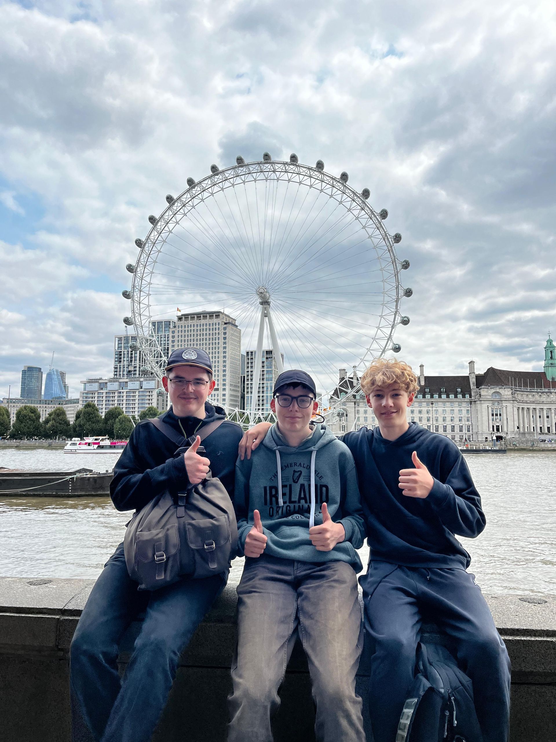 Drei Personen zeigen vor dem Riesenrad London Eye den Daumen nach oben.