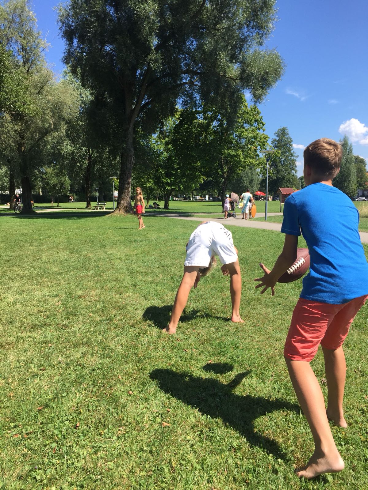 Drei barfüßige Personen spielen in einem sonnigen Park mit einem Fußball. Eine Person bückt sich, zwei andere machen sich bereit, ihn zu fangen.
