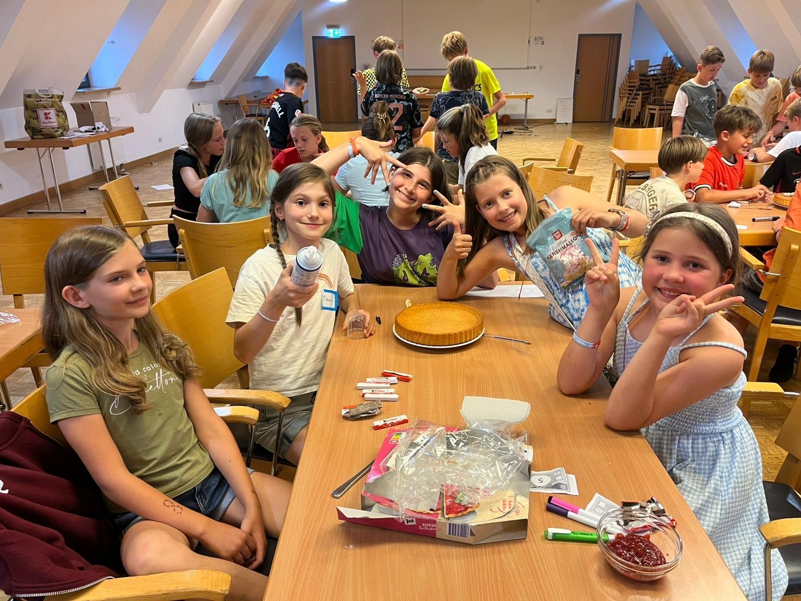 Kinder sitzen lächelnd an einem Tisch, davor steht ein Kuchen. Im Hintergrund sind weitere Personen zu sehen. Innenaufnahme.