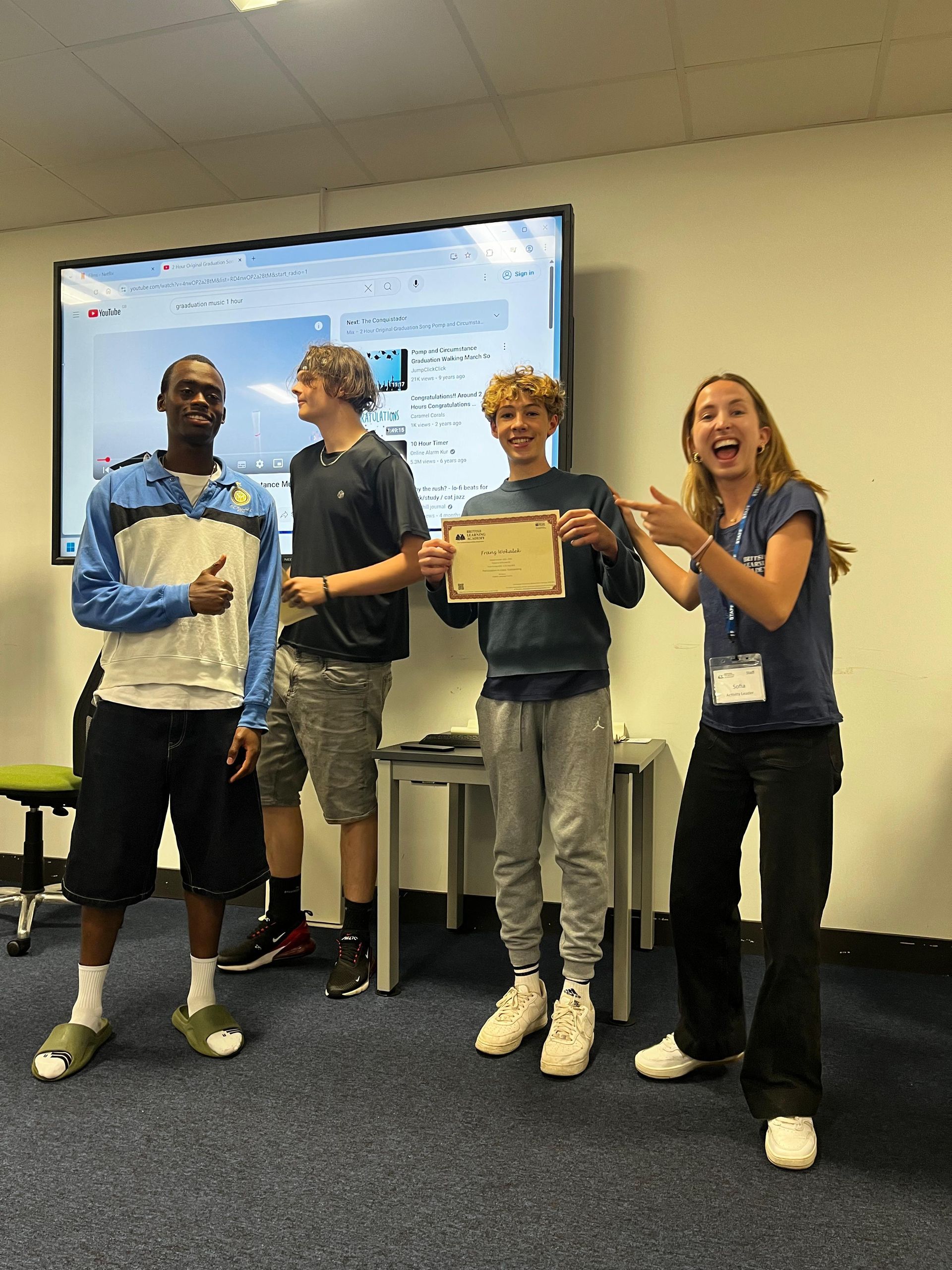 Vier Personen feiern; eine hält eine Urkunde. Raum mit Leinwand und blauem Teppich.