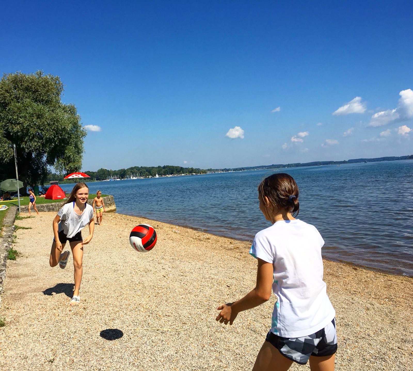 Zwei Personen spielen an einem sonnigen Tag Beachvolleyball an einem See.