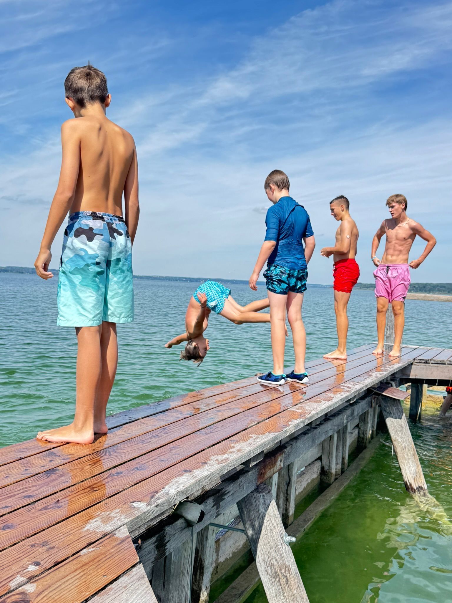Fünf Personen auf einem Holzsteg; eine macht einen Salto ins Wasser. Blauer Himmel und Wasser.