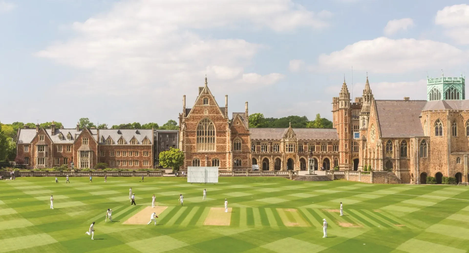 Cricketspiel auf einem gepflegten grünen Feld vor einem großen Backsteingebäude im gotischen Stil an einem sonnigen Tag.