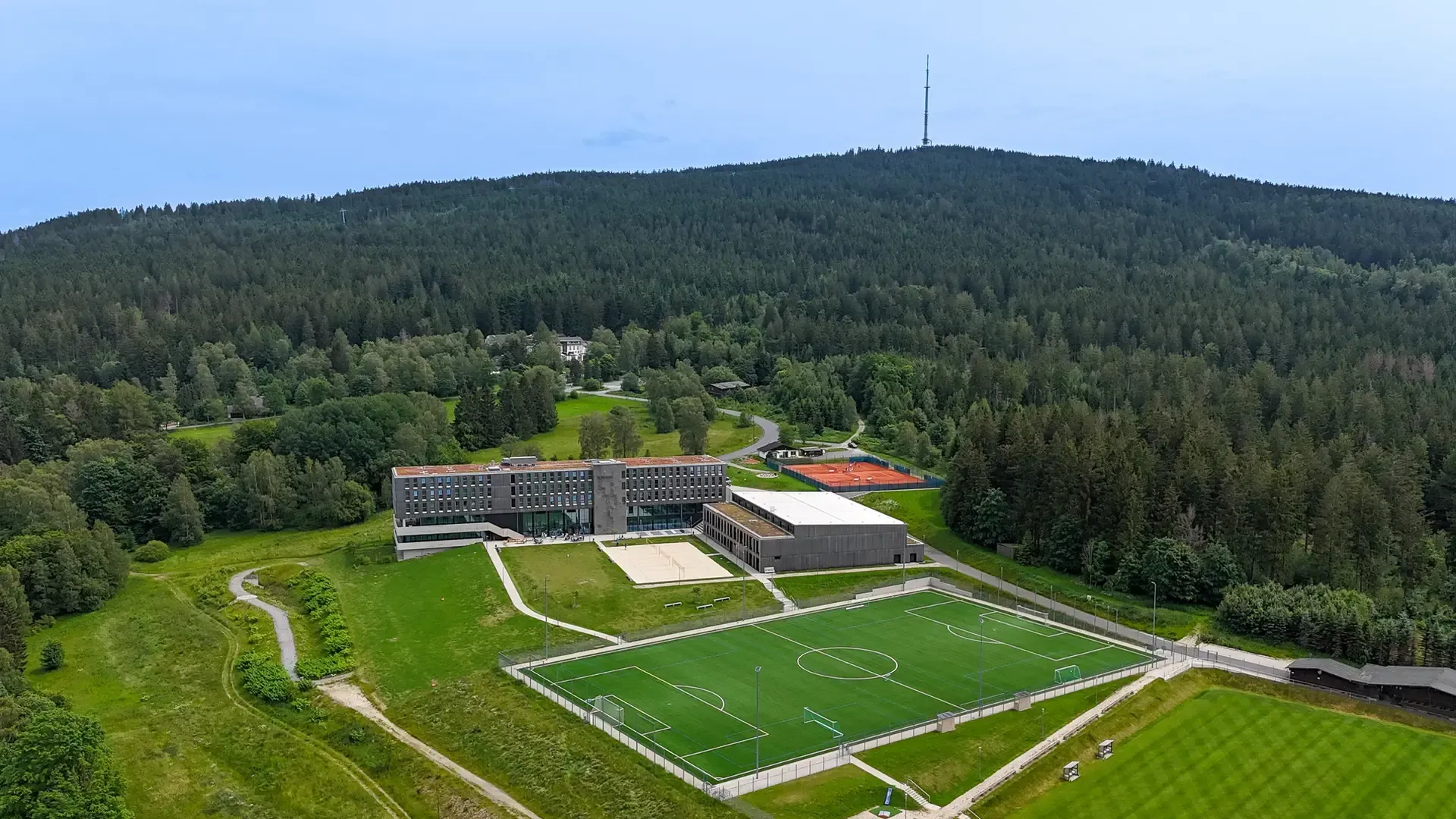 Ein Hotel und ein Sportplatz am Fuße eines bewaldeten Berges, auf dem ein Kommunikationsturm thront.