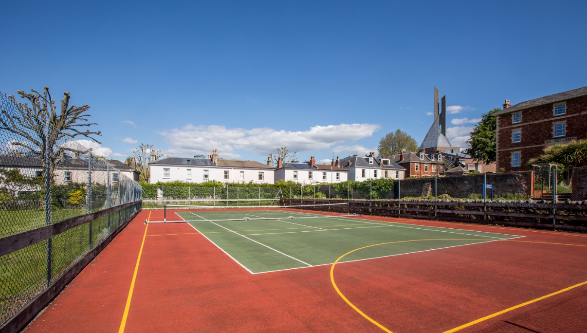 Freiluft-Sportplatz mit roten und grünen Spielflächen, Zaun, Gebäuden und blauem Himmel.