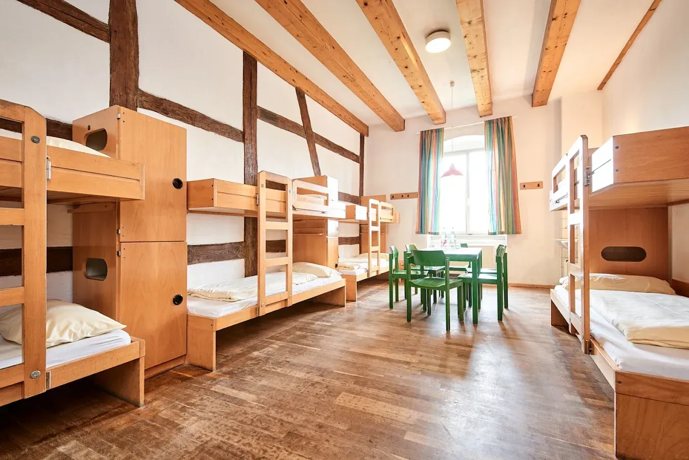 Schlafsaal mit mehreren hölzernen Etagenbetten, Holzbalken, einem kleinen Tisch mit Stühlen und einem Fenster mit grünen Vorhängen.