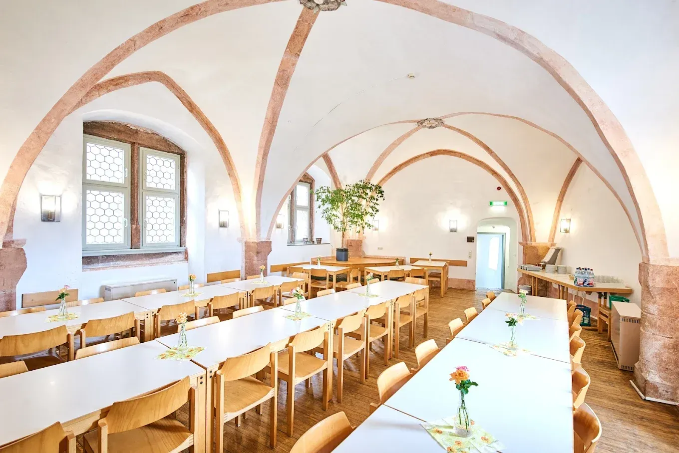 Ein langer Raum mit gewölbter Decke und Tischreihen, gedeckt für eine Veranstaltung. Sonnenlicht strömt durch ein großes Fenster herein.