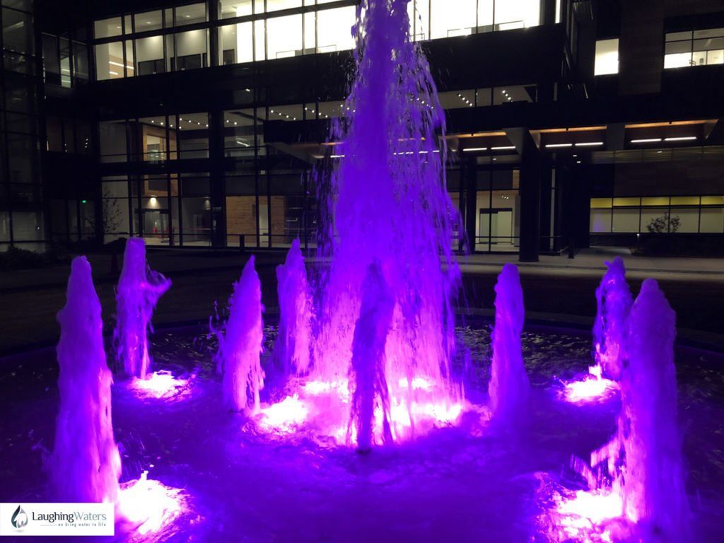 A purple fountain is lit up at night in front of a building.