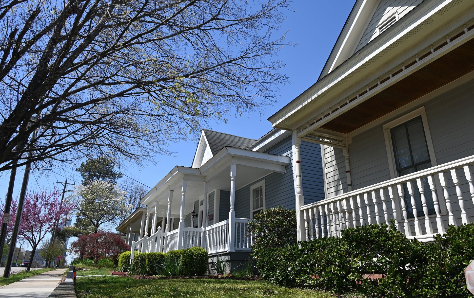 Row of white houses with porches along a sunny suburban street, with bare trees and green shrubs