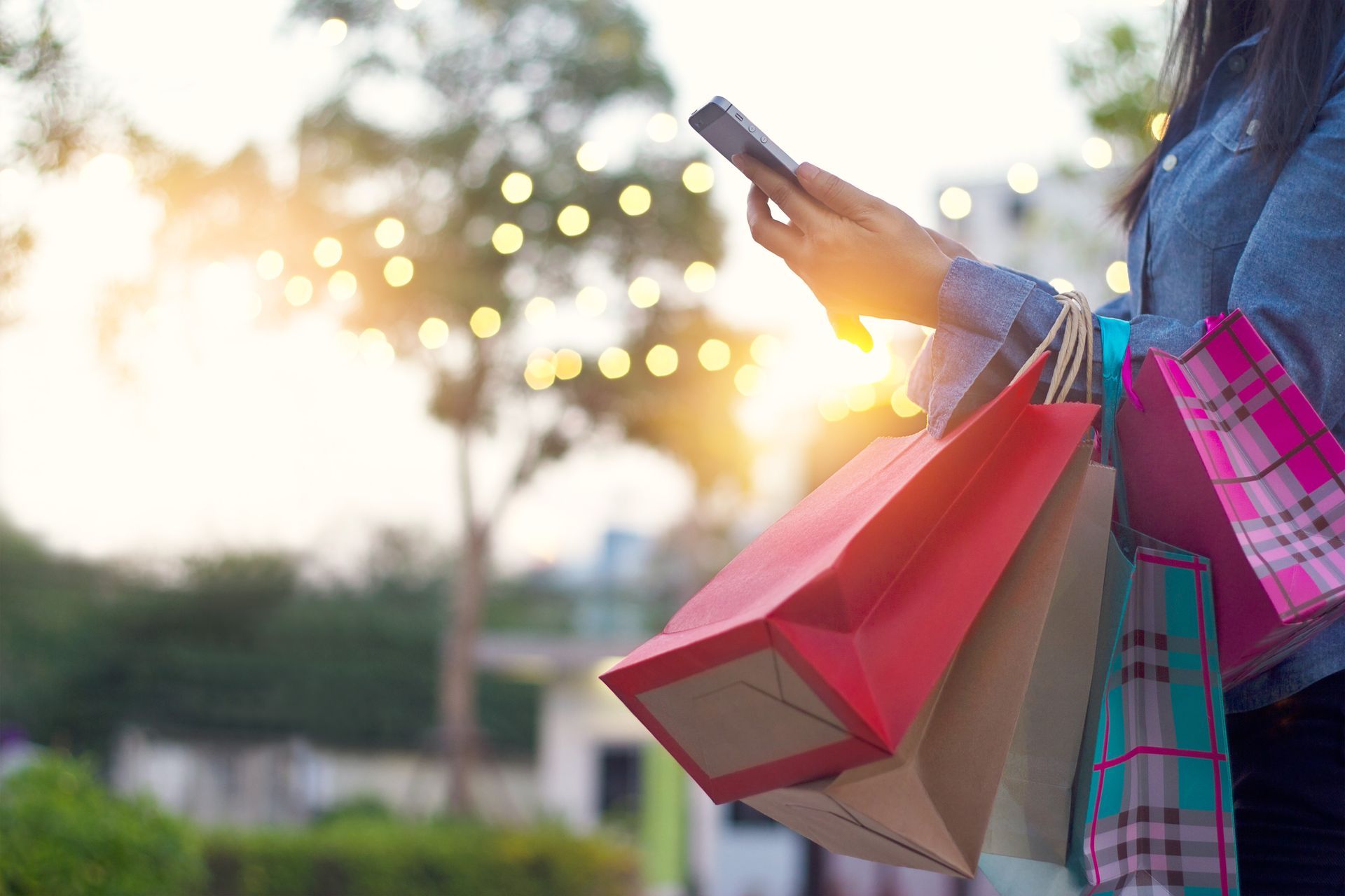 A close-up of a person holding a smartphone in one hand and carrying red and brown paper shopping bags in the other. The background features a soft, blurred outdoor setting with a tree and warm bokeh lights from string lamps glowing at sunset.