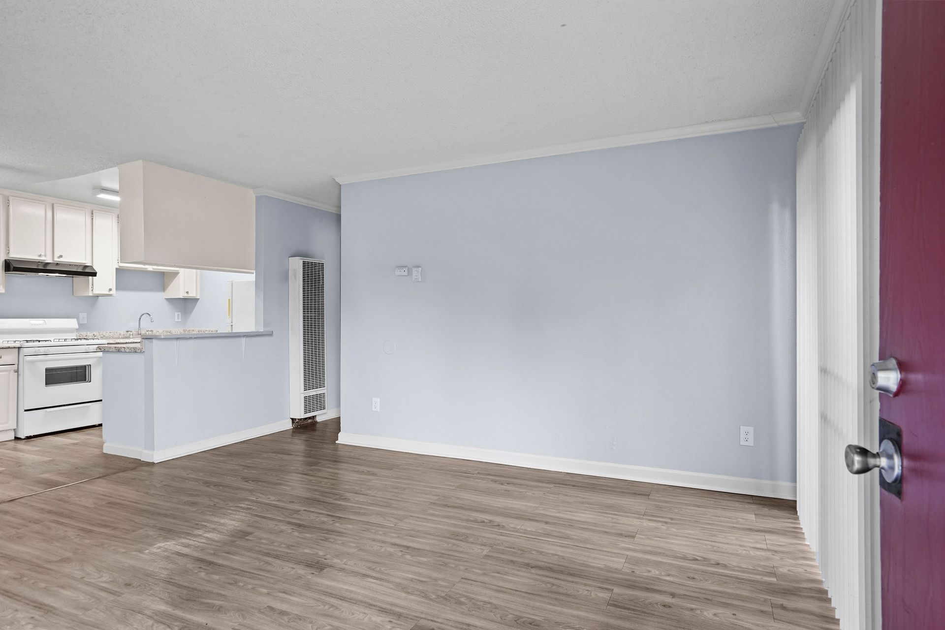 Empty living room with light gray walls, wood floors, and a view into a white kitchen