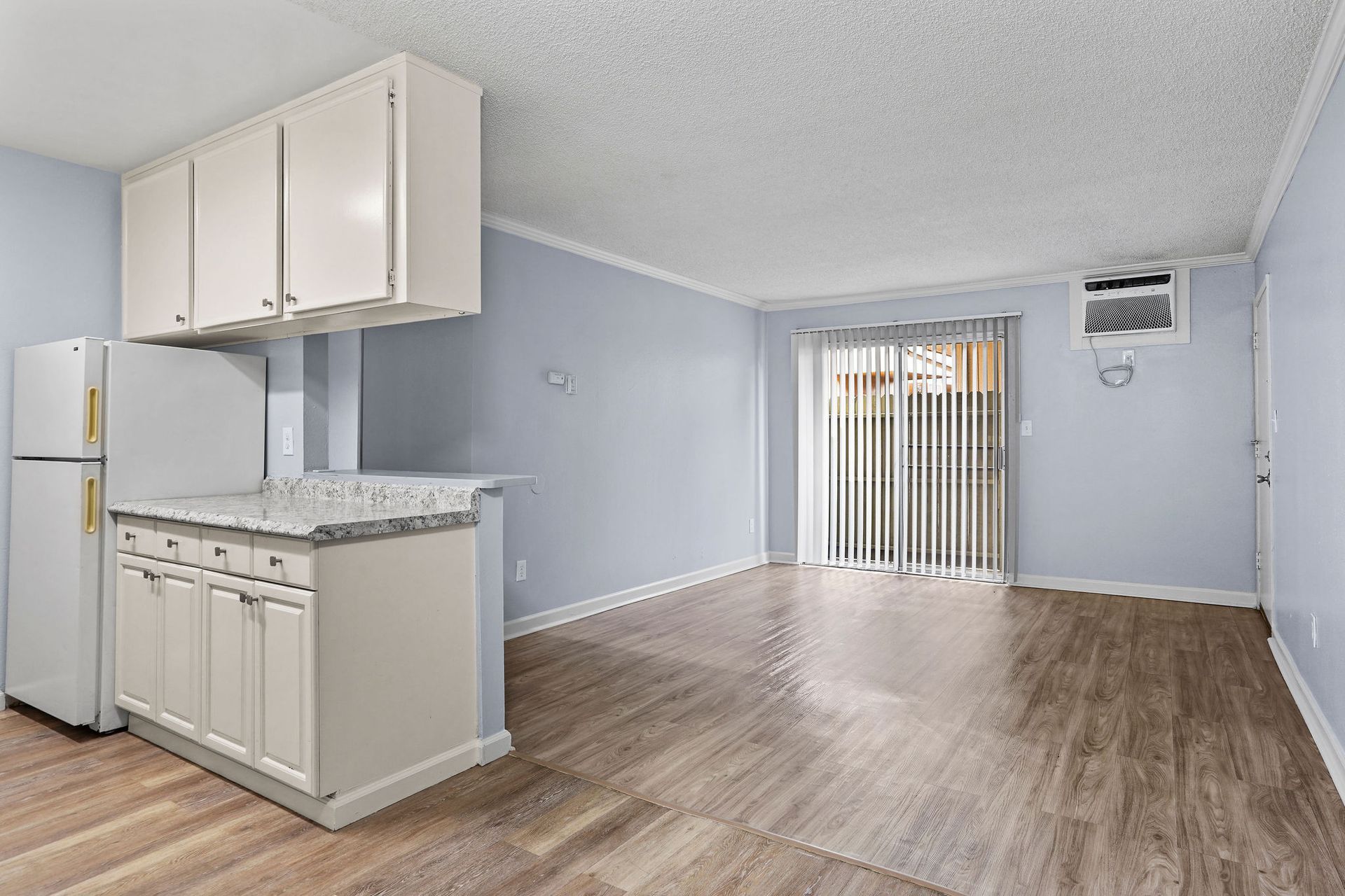 Empty apartment kitchen and living area with light gray walls, wood floors, and sliding glass doors.