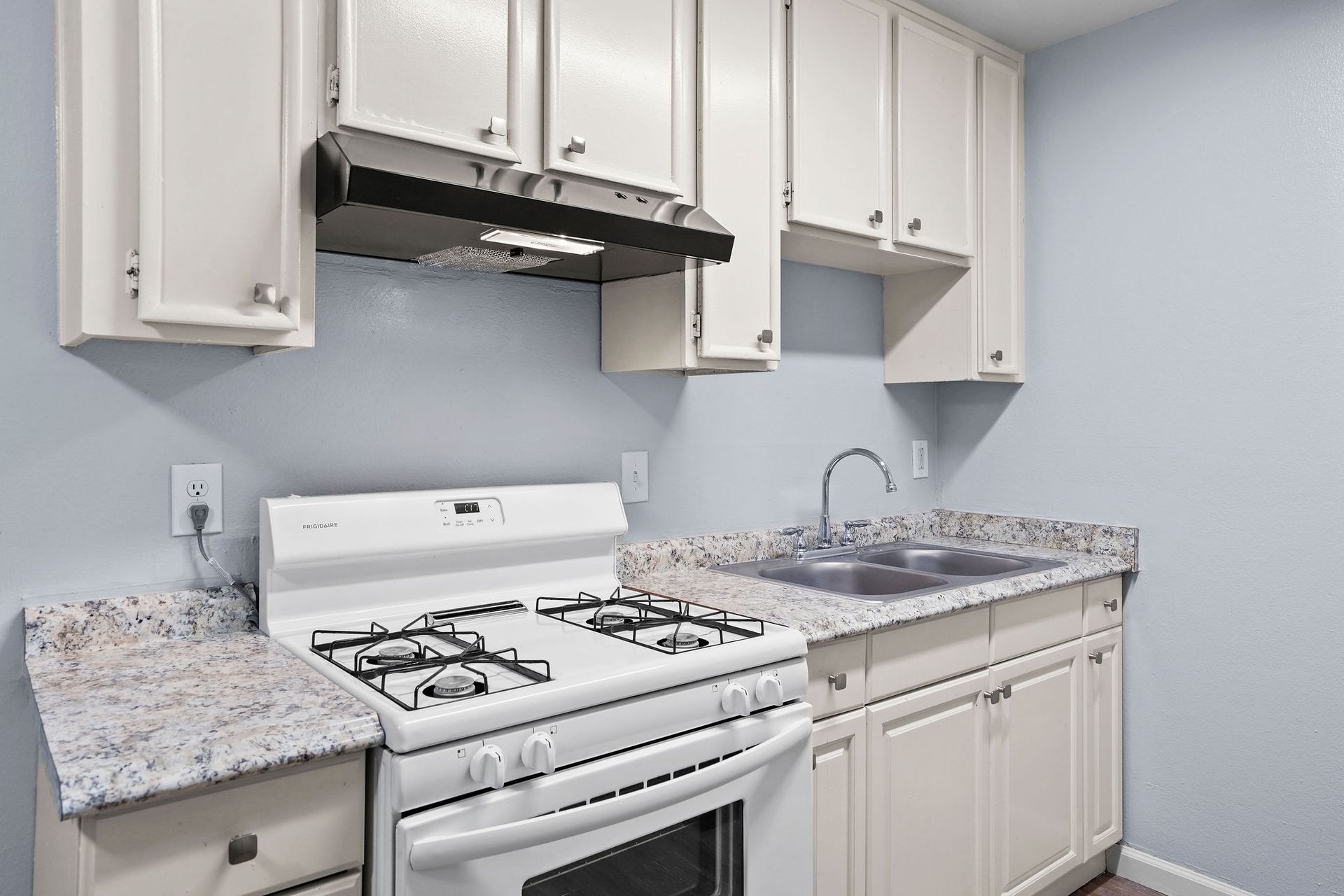 Small white kitchen with gas stove, speckled countertops, and white cabinets against a light gray wall.