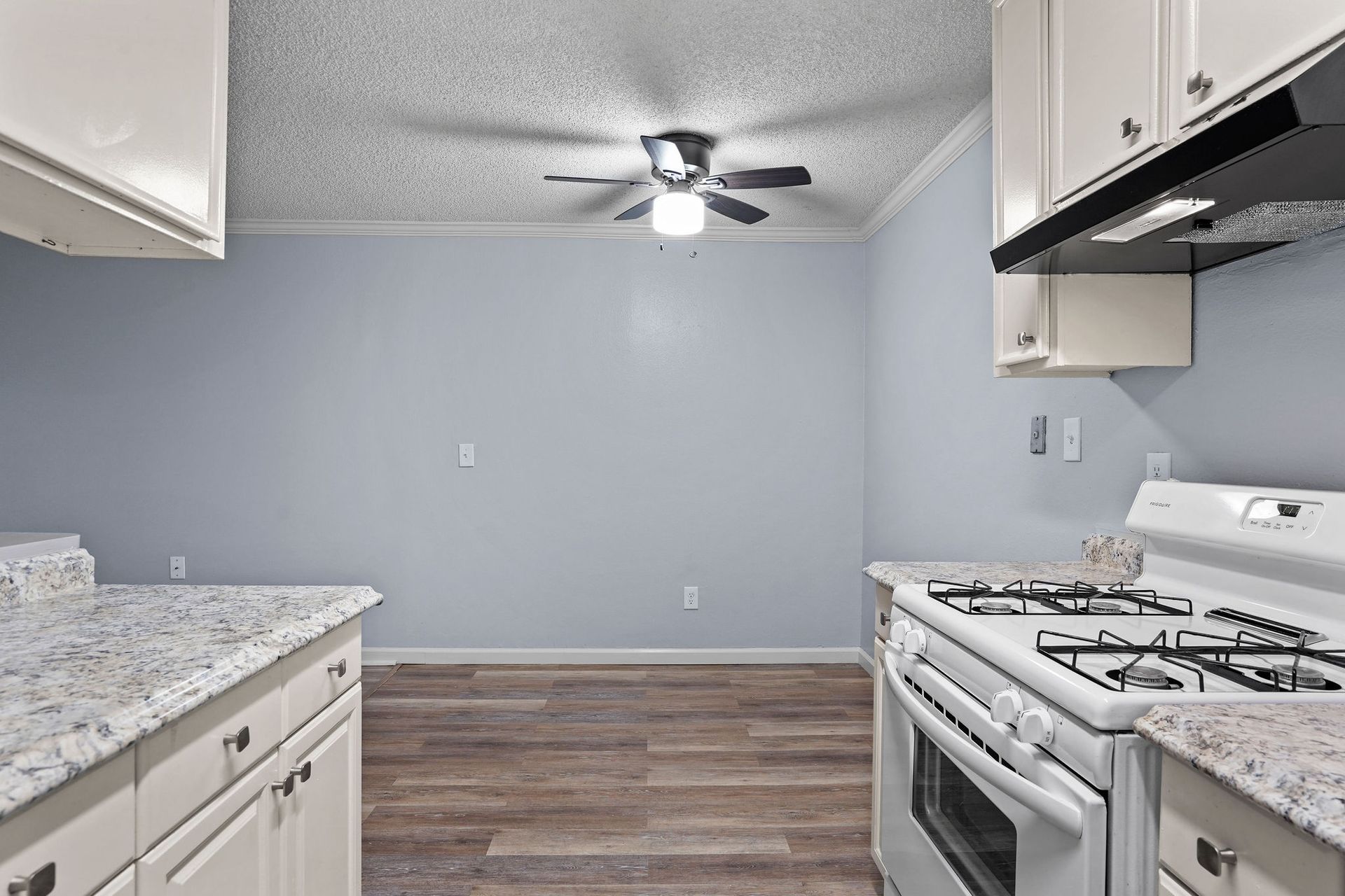 Empty kitchen with gray walls, white cabinets, granite counters, wood-look flooring, and a ceiling fan.