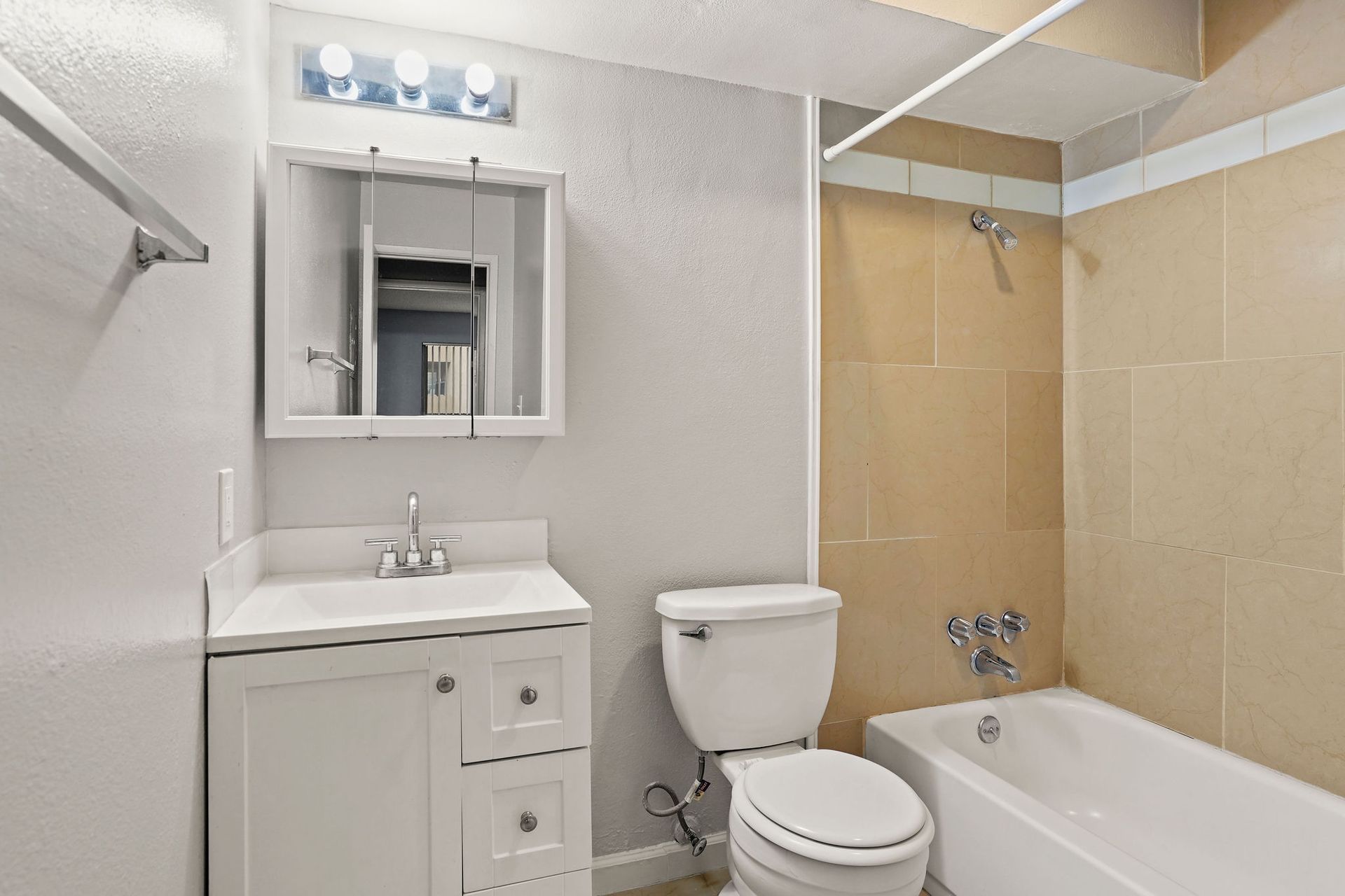 Small bathroom with white vanity, toilet, and bathtub beside beige tiled walls.