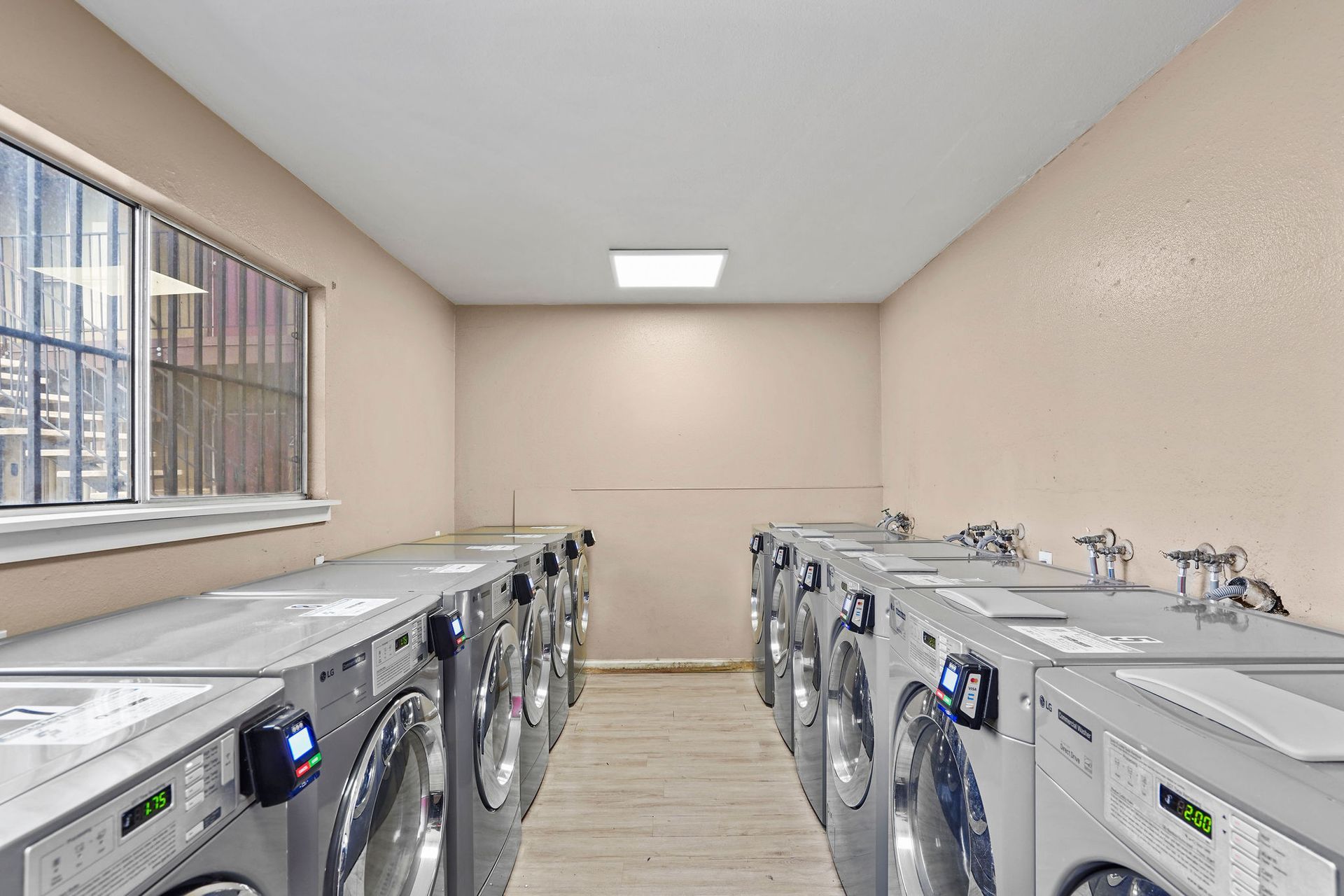 Rows of silver washing machines in a bright laundry room with a window and beige walls