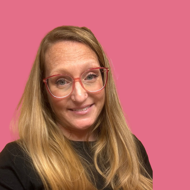 Woman with long blonde hair, wearing red-framed glasses, smiles against a pink backdrop.