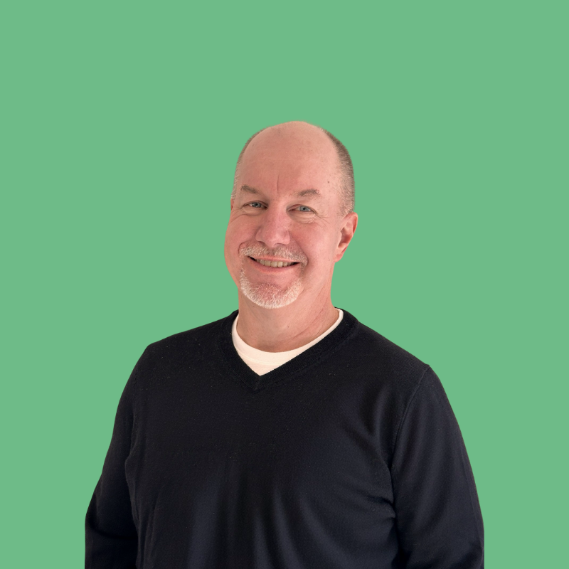 Smiling man with a shaved head and graying goatee in a black sweater against a green background.