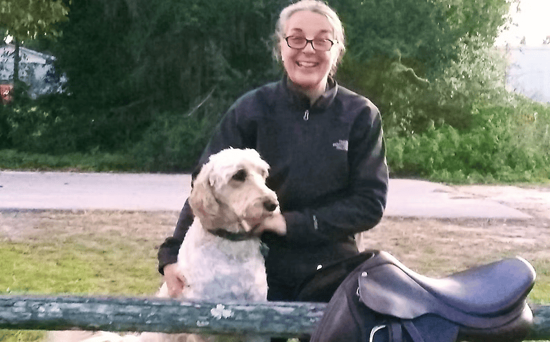 Woman in black jacket with a light-colored dog, smiling, beside a saddle, outdoors.